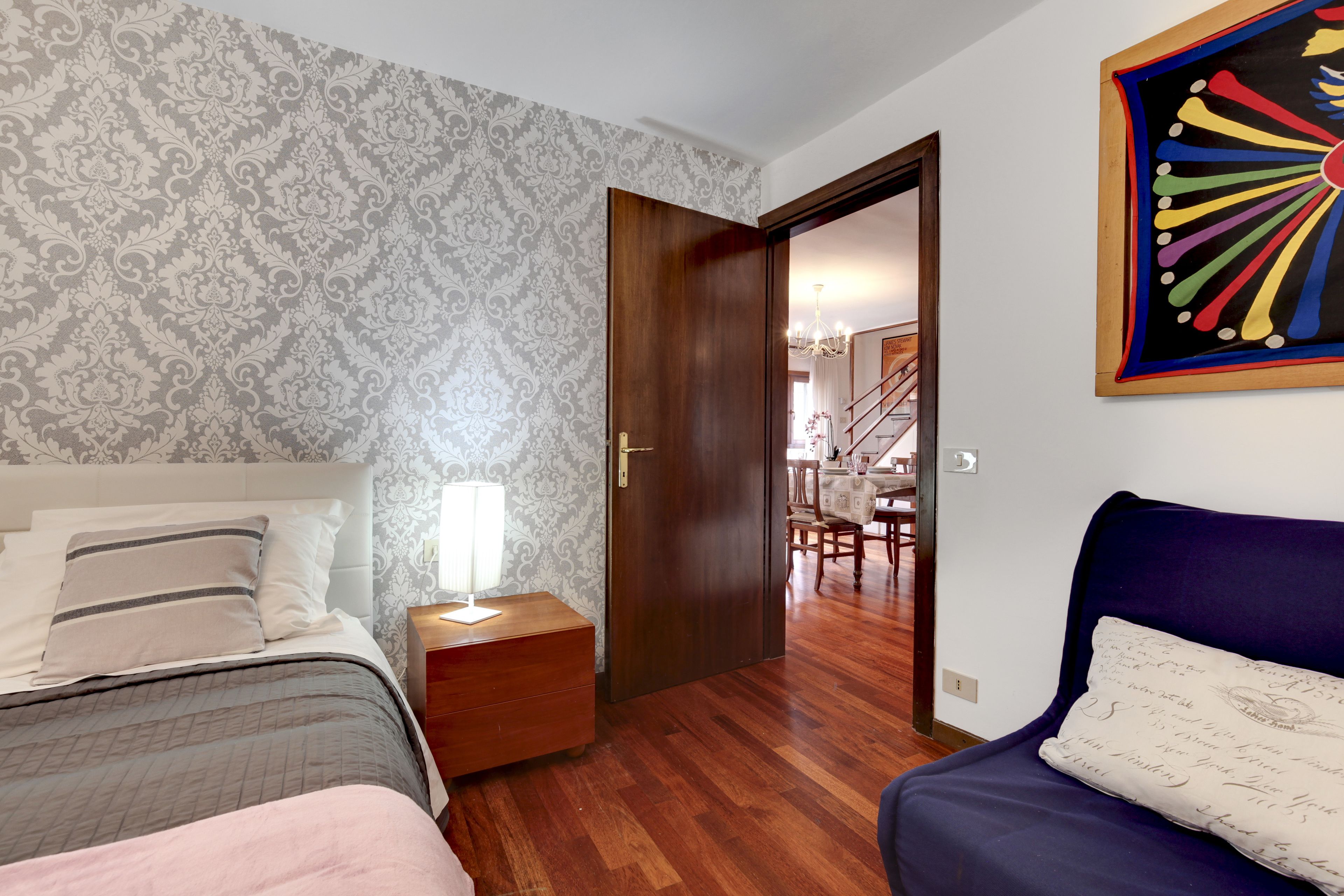 A bedroom with a bed, nightstand, dresser, and a door leading to a dining area.