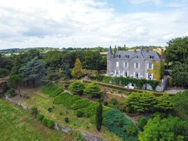 Um edifício grande e histórico com fachada simétrica, cercado por vegetação exuberante e um jardim bem cuidado.