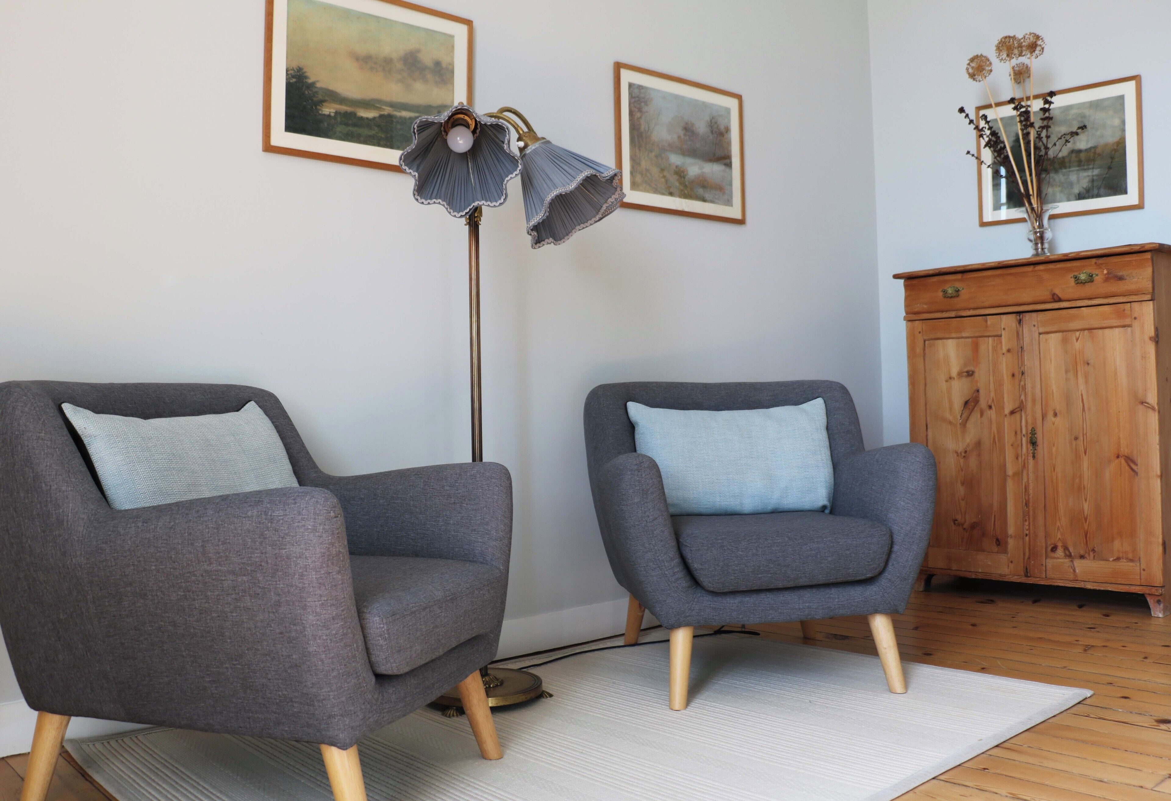 Two grey armchairs with blue cushions, a tall floor lamp, and a wooden cabinet with framed pictures on the wall.