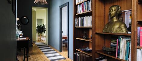 A hallway with wooden flooring, a bookshelf with books, a bust sculpture, and a hanging light fixture.