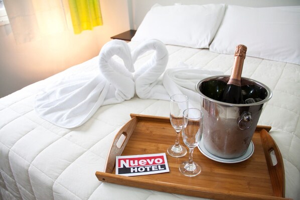 A bed with white linens, a heart-shaped towel arrangement, a bottle of champagne, two glasses, and a wooden tray with a hotel sign. - Nuevo Hotel (Rivera)