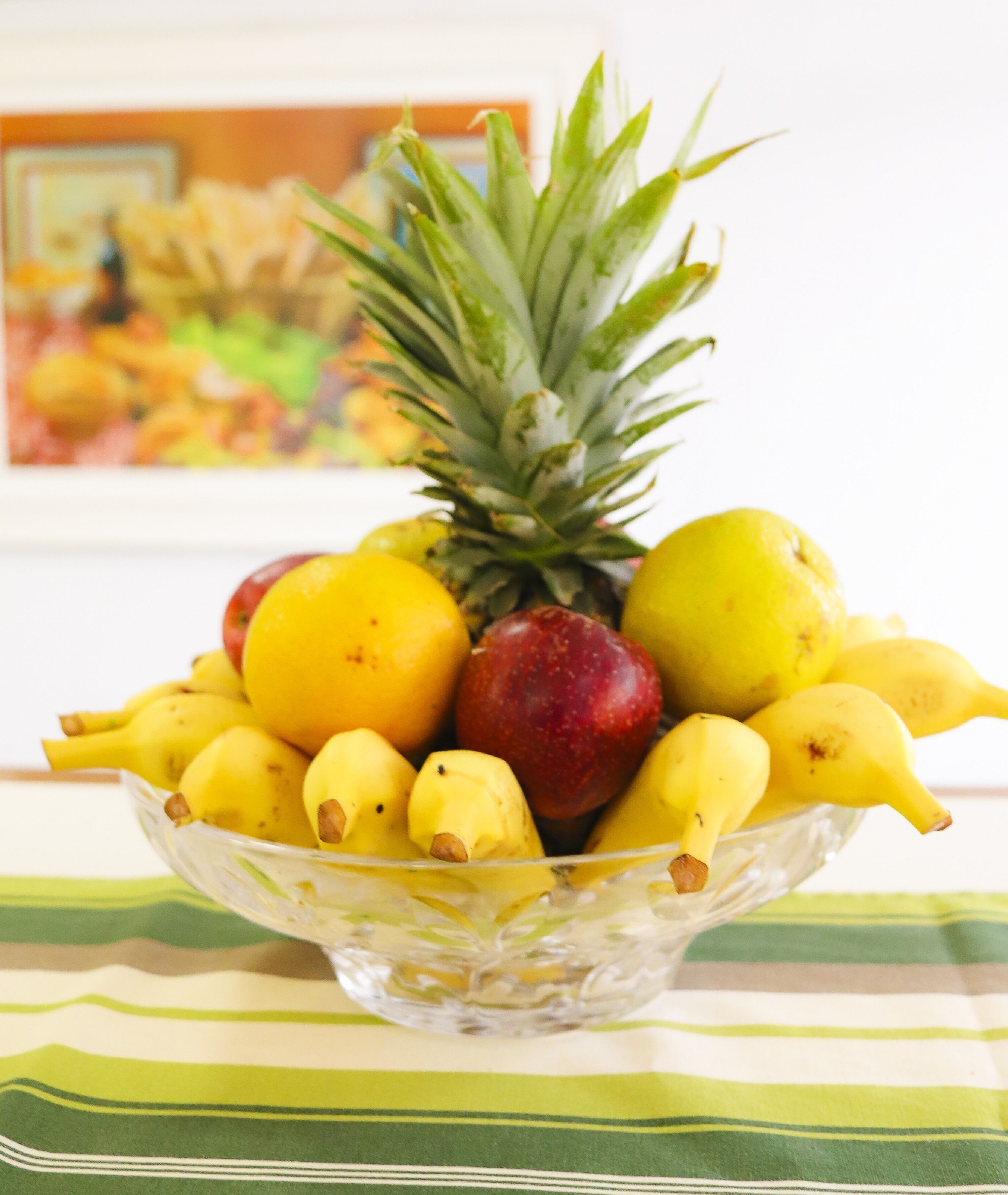 Un saladier en verre rempli de bananes, de pommes et d’un ananas, posé sur une nappe à rayures.