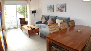 Un salon moderne avec un canapé gris, une table basse en bois et une table à manger avec des chaises. On y trouve deux tableaux accrochés au mur et une lampe sur pied.