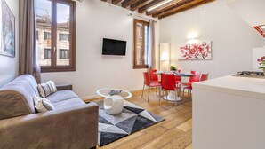 A modern living room with a sofa, a coffee table, and a dining area with red chairs and a white table. - Residence Le Beccarie - Rialto by Wonderful Italy (Venice)