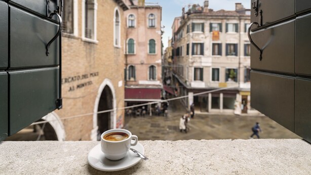 A cup of coffee on a table with a view of a street scene through an open window. - Residence Le Beccarie - Rialto by Wonderful Italy (Venice)