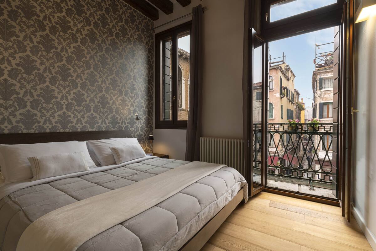 A bedroom with a large bed, a window with a view of buildings, and a balcony with a railing.