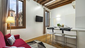 A modern living room with a red sofa, a patterned rug, a TV mounted on the wall, and a kitchen area with white bar stools. - Residence Le Beccarie - Rialto by Wonderful Italy (Venice)