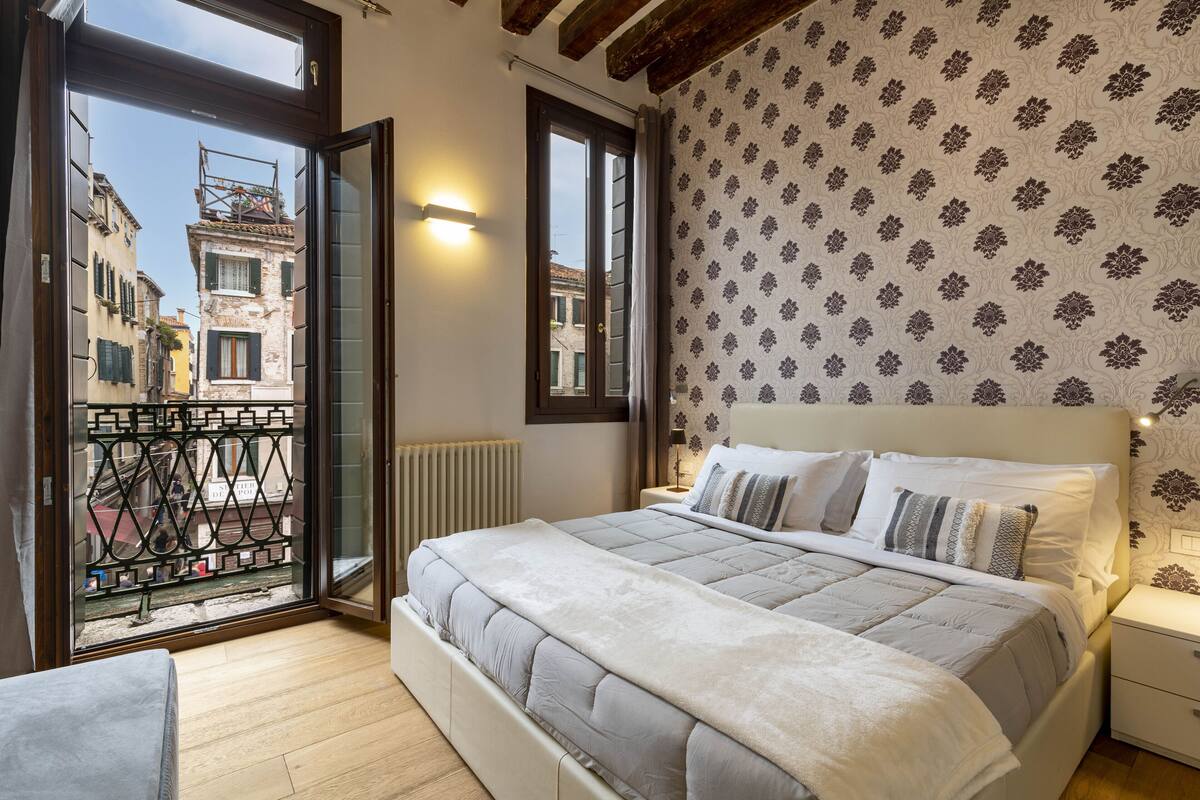 A bedroom with a bed, a nightstand, a window with a view of buildings, and a balcony with a railing.