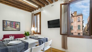 A dining area with a table set for two, a television mounted on the wall, and a window with a view of a street. - Residence Le Beccarie - Rialto by Wonderful Italy (Venice)