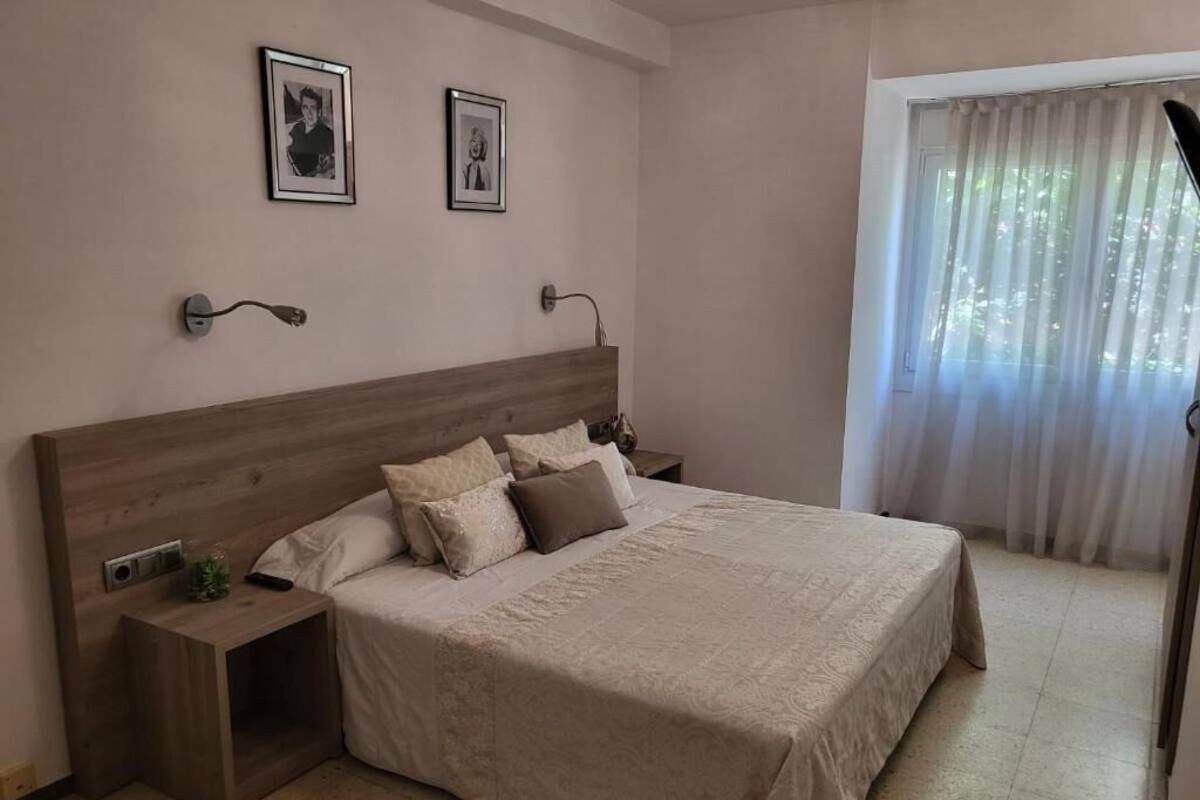 A bedroom with a wooden headboard, a bed with beige bedding, two bedside tables, and a window with sheer curtains.