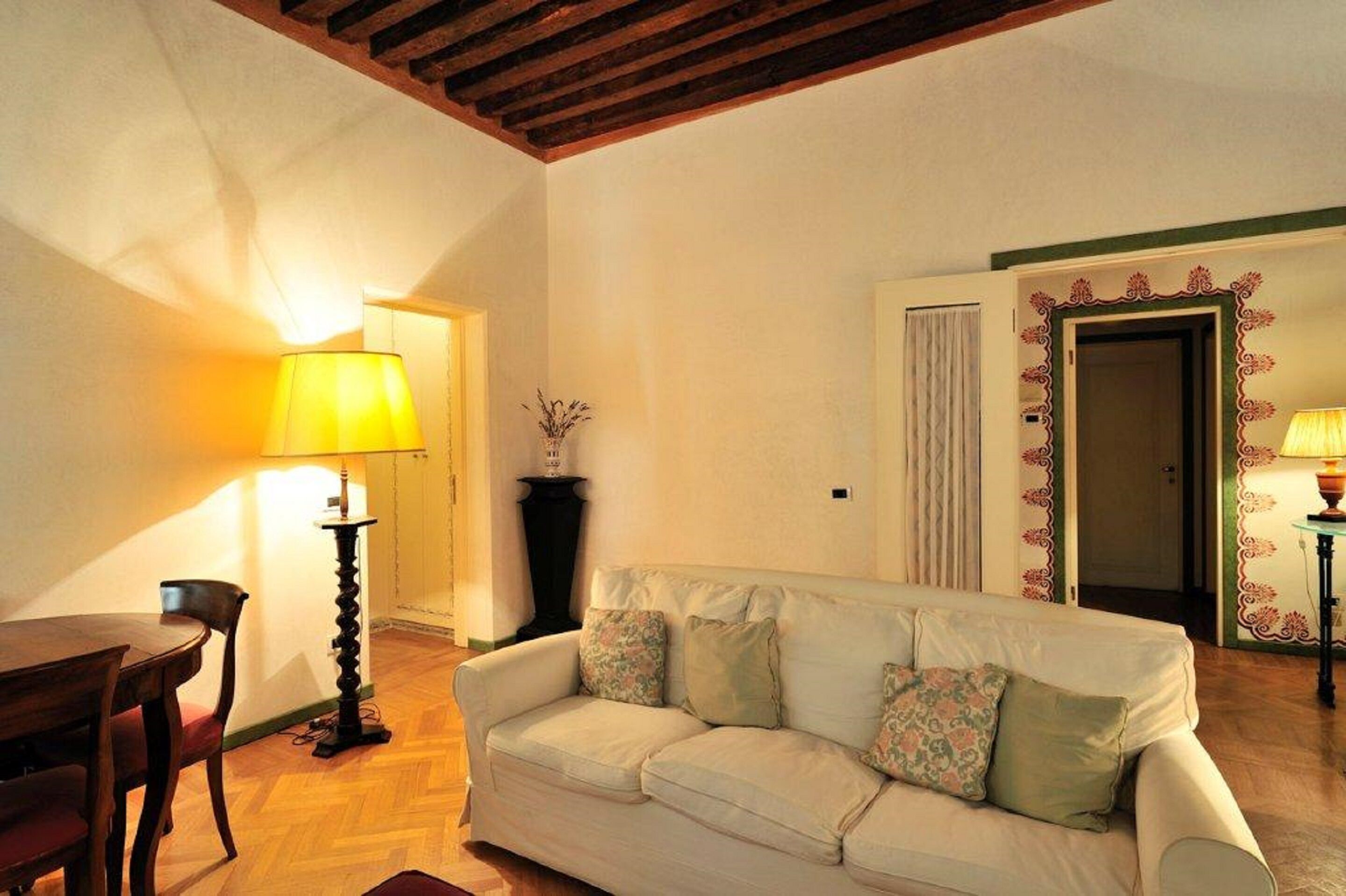 Una sala de estar con techo de madera, un sofá blanco con cojines estampados, una mesa de comedor de madera y una lámpara.