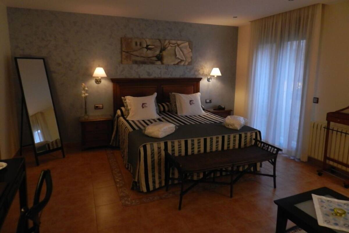 Une chambre d’hôtel avec un grand lit, deux lampes de chevet, un miroir, un banc et une petite table sur laquelle se trouve un livre.
