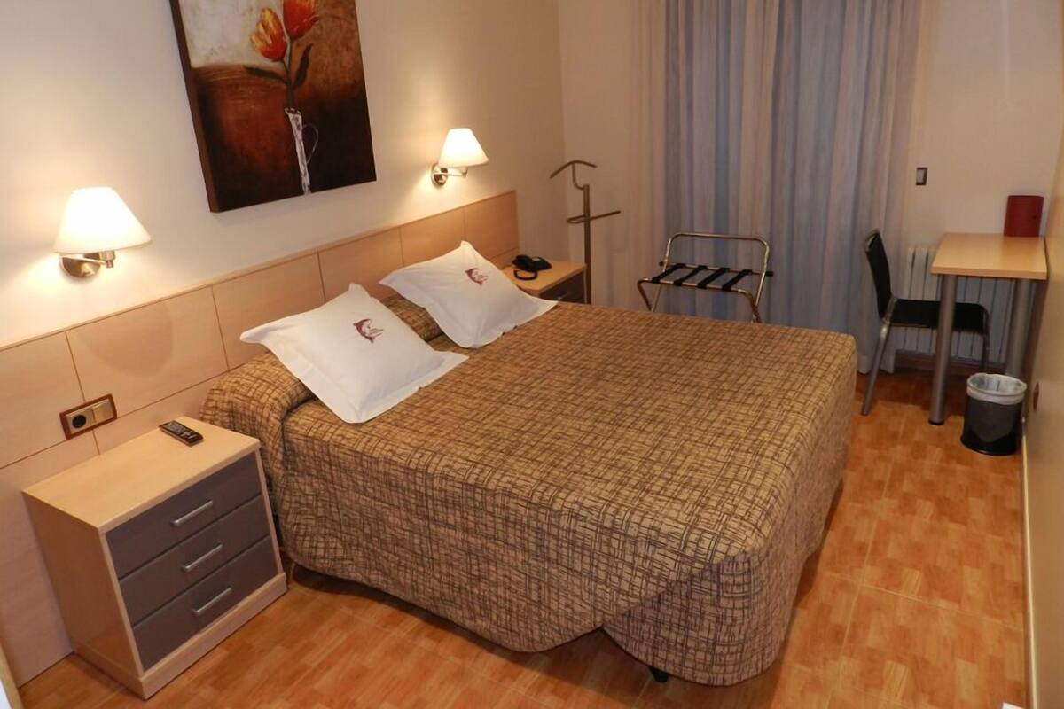 Une chambre d’hôtel comprenant un lit, des tables de chevet, un bureau, une chaise, une lampe, un tableau et une poubelle.