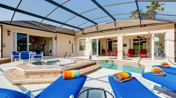 A covered outdoor pool area with blue lounge chairs, a hot tub, and a patio with red chairs.