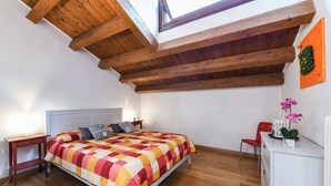 Ein Schlafzimmer mit Holzbalkendecke, einem Bett mit kariertem Überwurf, einem Nachttisch, einem Sessel und einer Kommode mit Blumen in einer Vase.