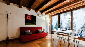 Ein modernes Wohnzimmer mit einem roten Sofa, Holzboden, einem Esstisch mit Stühlen und großen Fenstern mit Vorhängen.