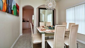 A dining area with a glass table, white chairs, and a chandelier. There is a colorful abstract painting on the wall and a kitchen in the background. - 616BD (Davenport)
