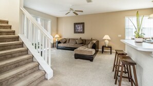 Un salon avec un canapé beige, un pouf et une table basse. Un escalier avec des rampes blanches mène à un autre niveau.