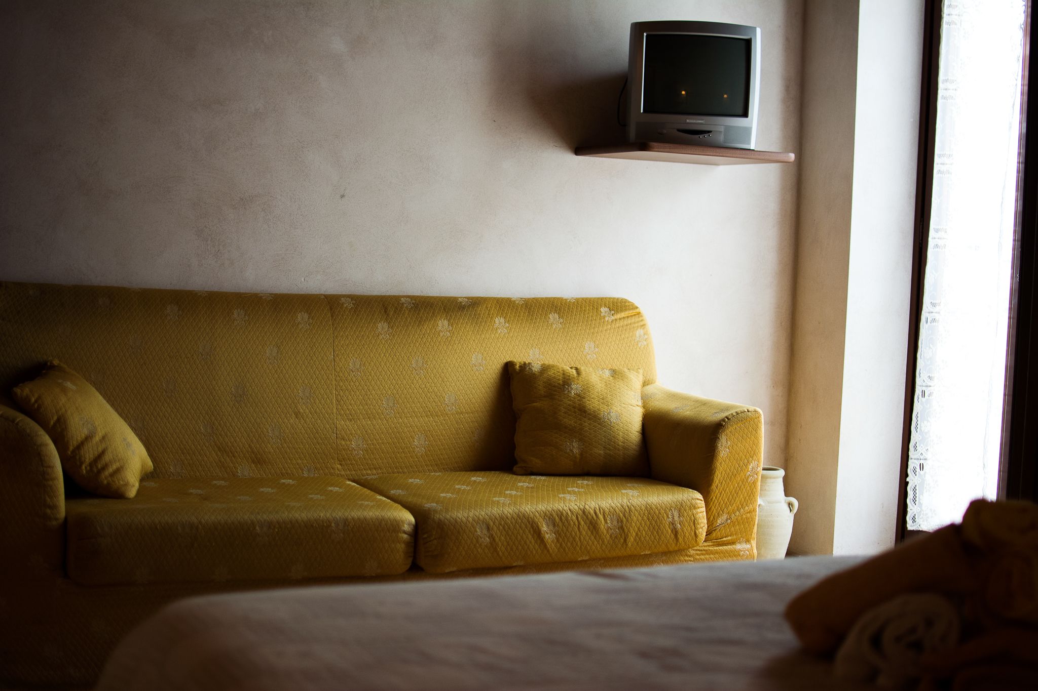 A yellow sofa with pillows, a television on a shelf, and a window with curtains.