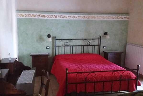 A bedroom with a metal bed frame, a red bedspread, a nightstand with a lamp, and a dresser.