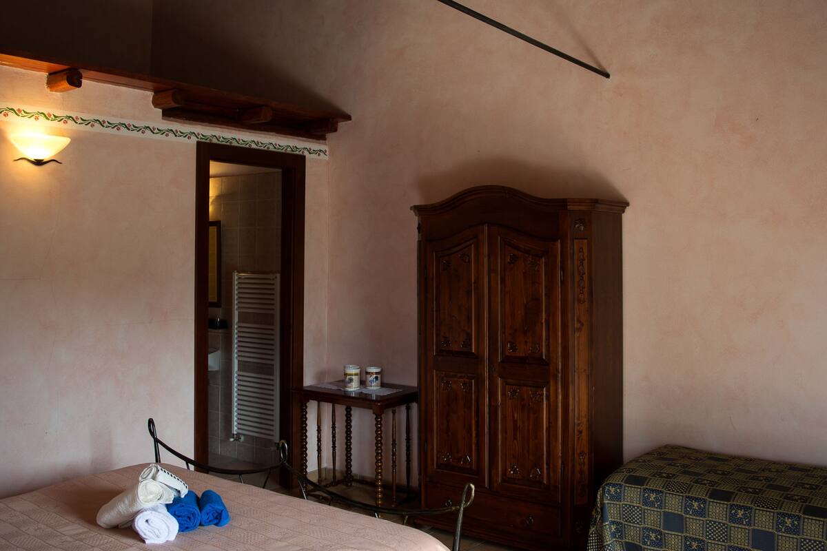 A room with a bed, a wooden wardrobe, a small table with items, and a wall-mounted light fixture.