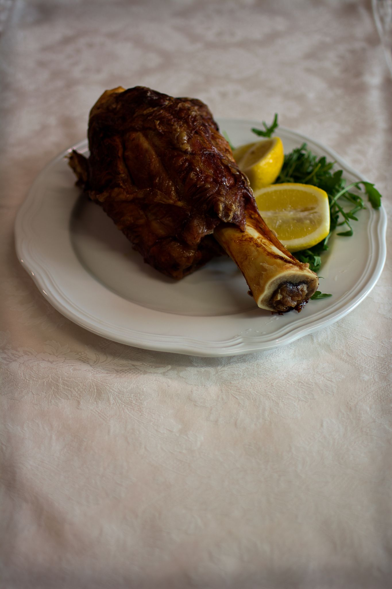 A roasted leg of lamb on a white plate with lemon and greens.