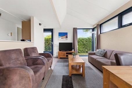A modern living room with a sofa, a coffee table, and a TV. There is a door leading to a balcony with a view of greenery.