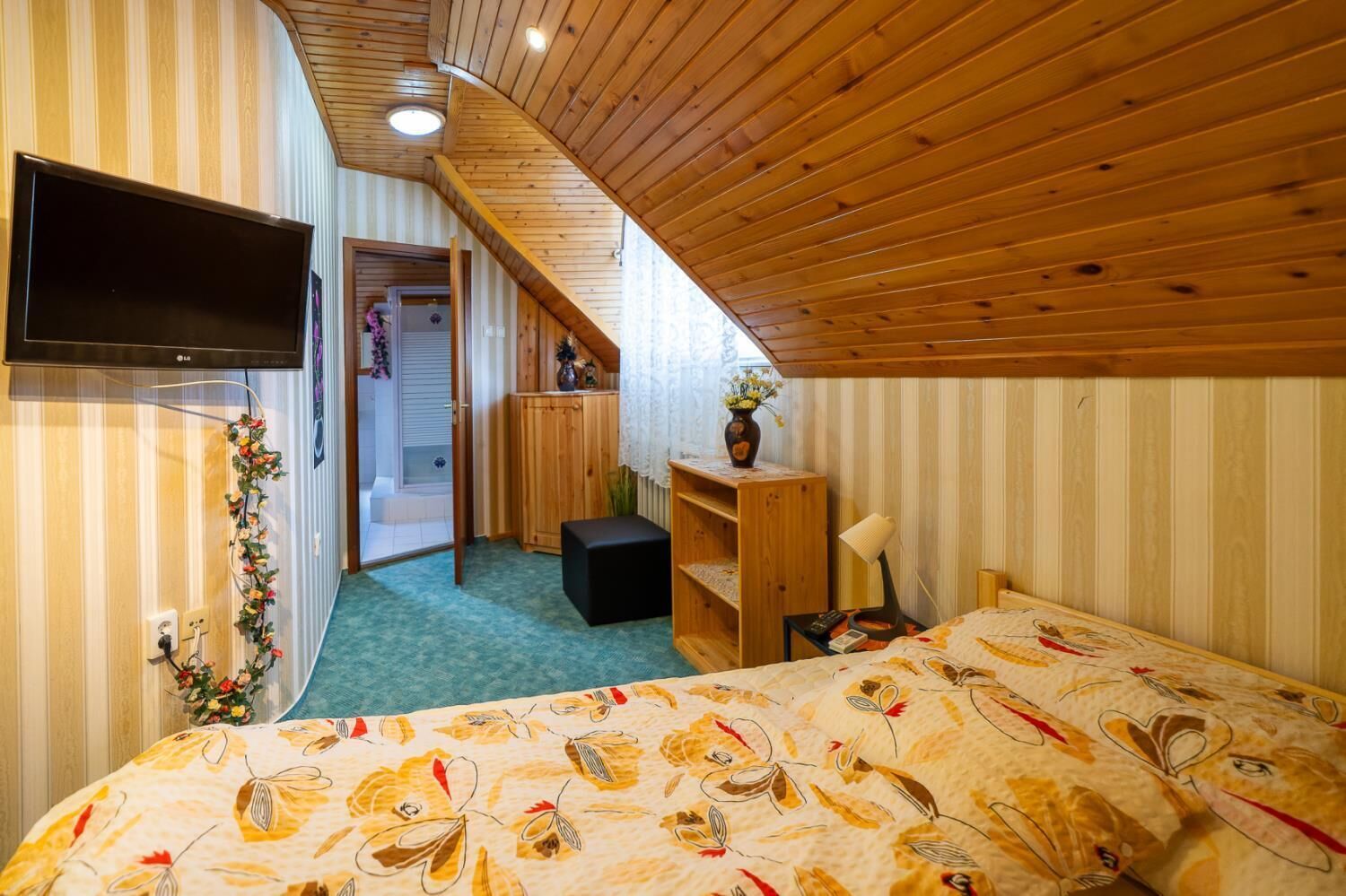 Een slaapkamer met schuine plafonds, een bed met een bloemenprint, een televisie, een klein houten kastje en een bureau met een lamp.