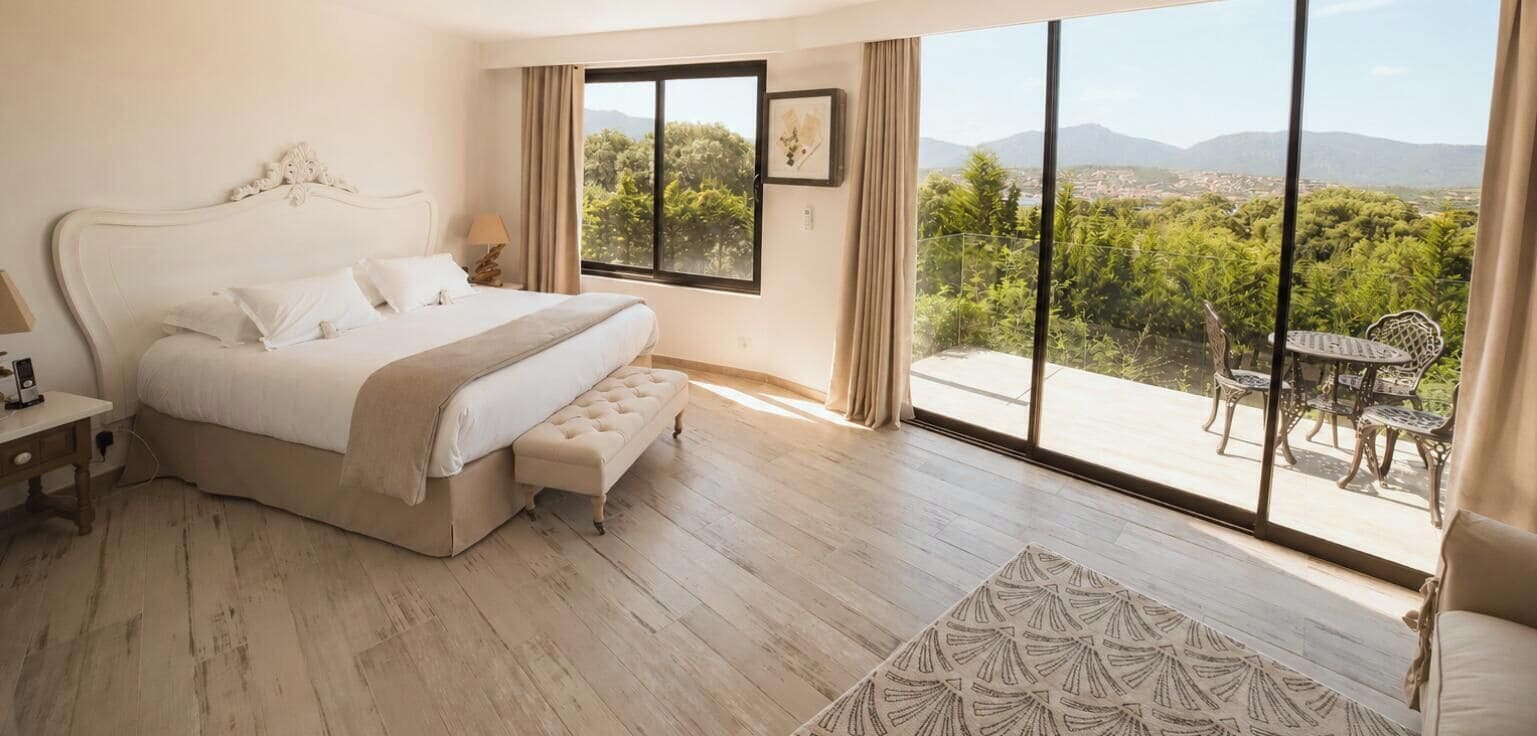 Ein Schlafzimmer mit einem großen Bett, einem Nachttisch, einem Fenster mit Blick auf Grünflächen und Berge sowie einem Balkon mit Tisch und Stühlen.