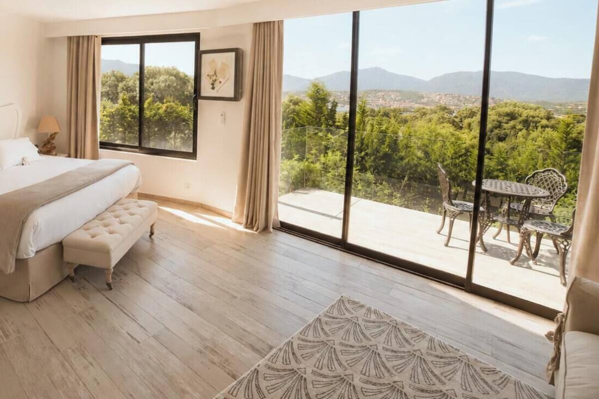 A bedroom with a large bed, a bedside table, a nightstand, a window with a view of greenery and mountains, and a balcony with outdoor furniture.