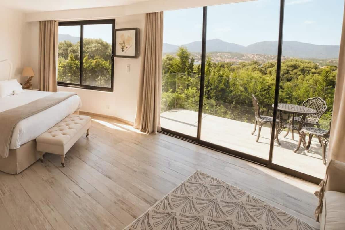 A bedroom with a large bed, a bedside table, a nightstand, a window with a view of greenery and mountains, and a balcony with a table and chairs.
