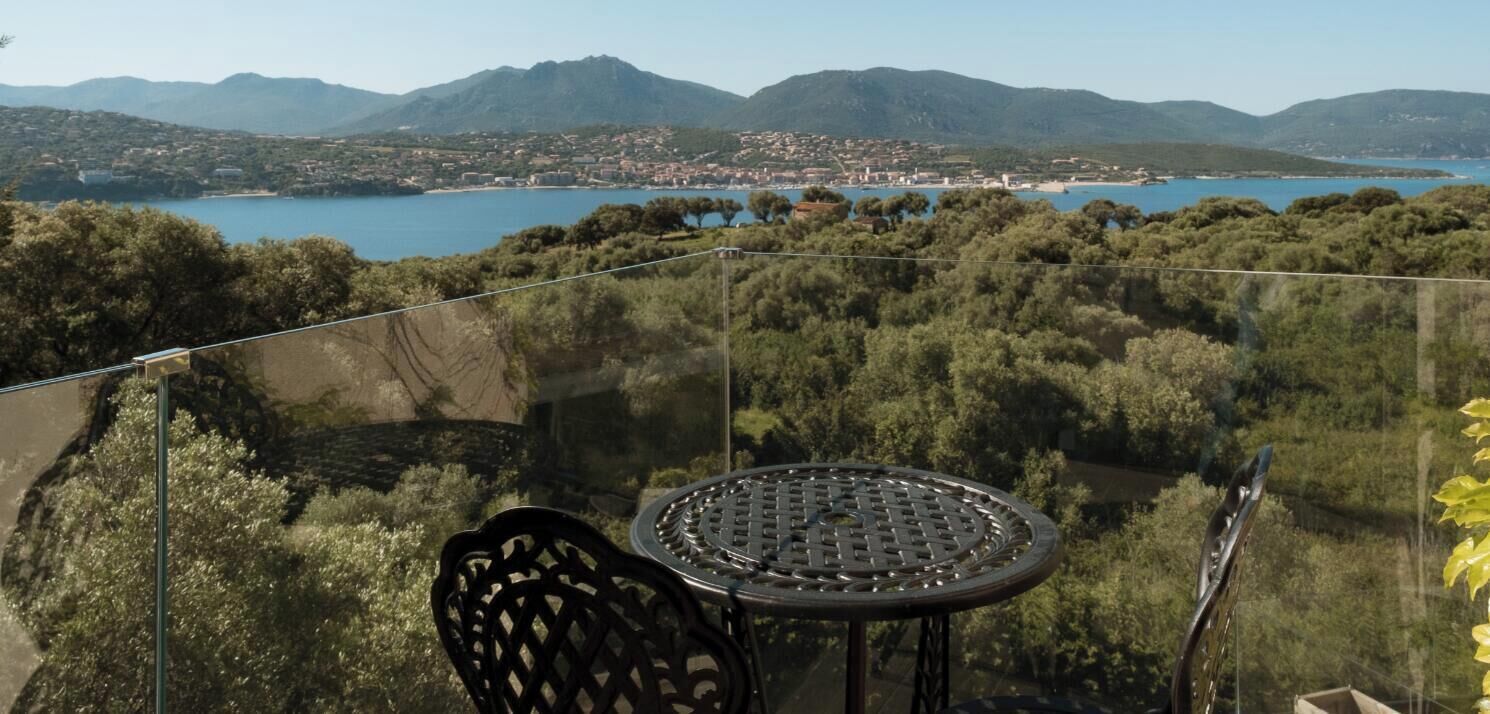 Un balcon avec une table et des chaises donnant sur un paysage côtier, avec des montagnes à l’horizon.