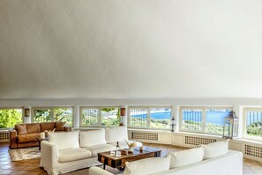 Uma sala de estar ampla com uma grande janela com vista para uma paisagem deslumbrante, um sofá marrom, um sofá branco e uma mesa de centro de madeira.