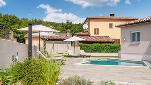 A swimming pool area with lounge chairs and umbrellas, surrounded by greenery and a building with a tiled roof. - Awesome home in Labin (Labin)