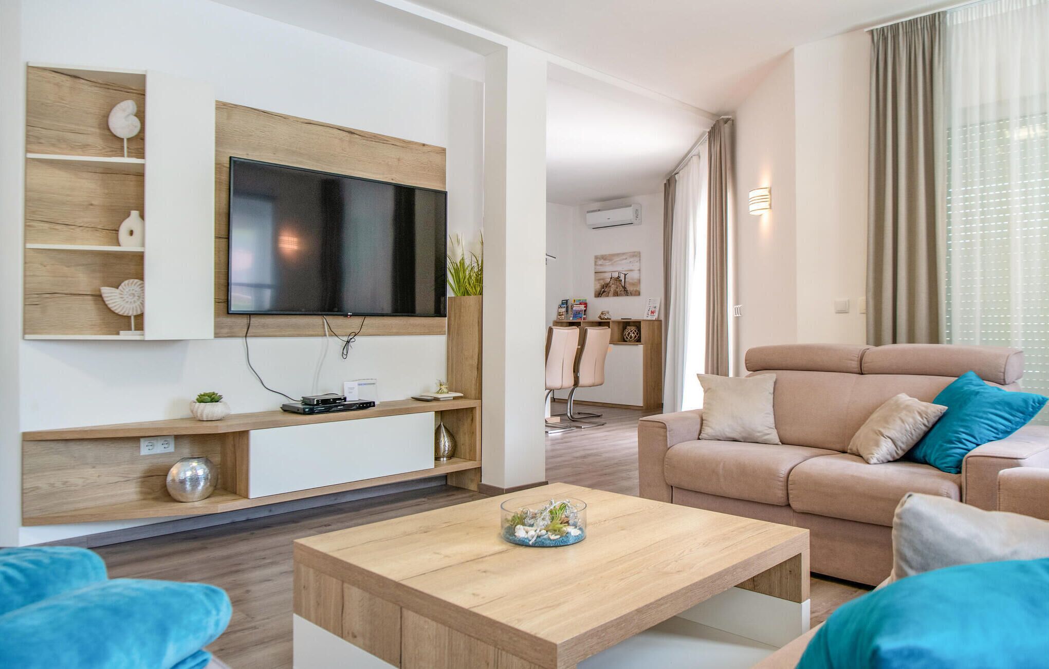 A modern living room with a flat-screen TV mounted on a wooden wall, a beige sofa with blue cushions, a wooden coffee table, and a dining area in the background.