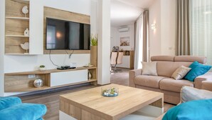 A modern living room with a flat-screen TV mounted on a wooden wall, a beige sofa with blue cushions, a wooden coffee table, and a dining area in the background. - Awesome home in Labin (Labin)