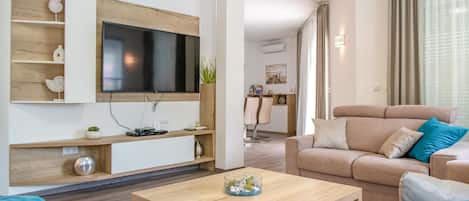 A modern living room with a flat-screen TV mounted on a wooden wall, a beige sofa with blue cushions, a wooden coffee table, and a dining area in the background.