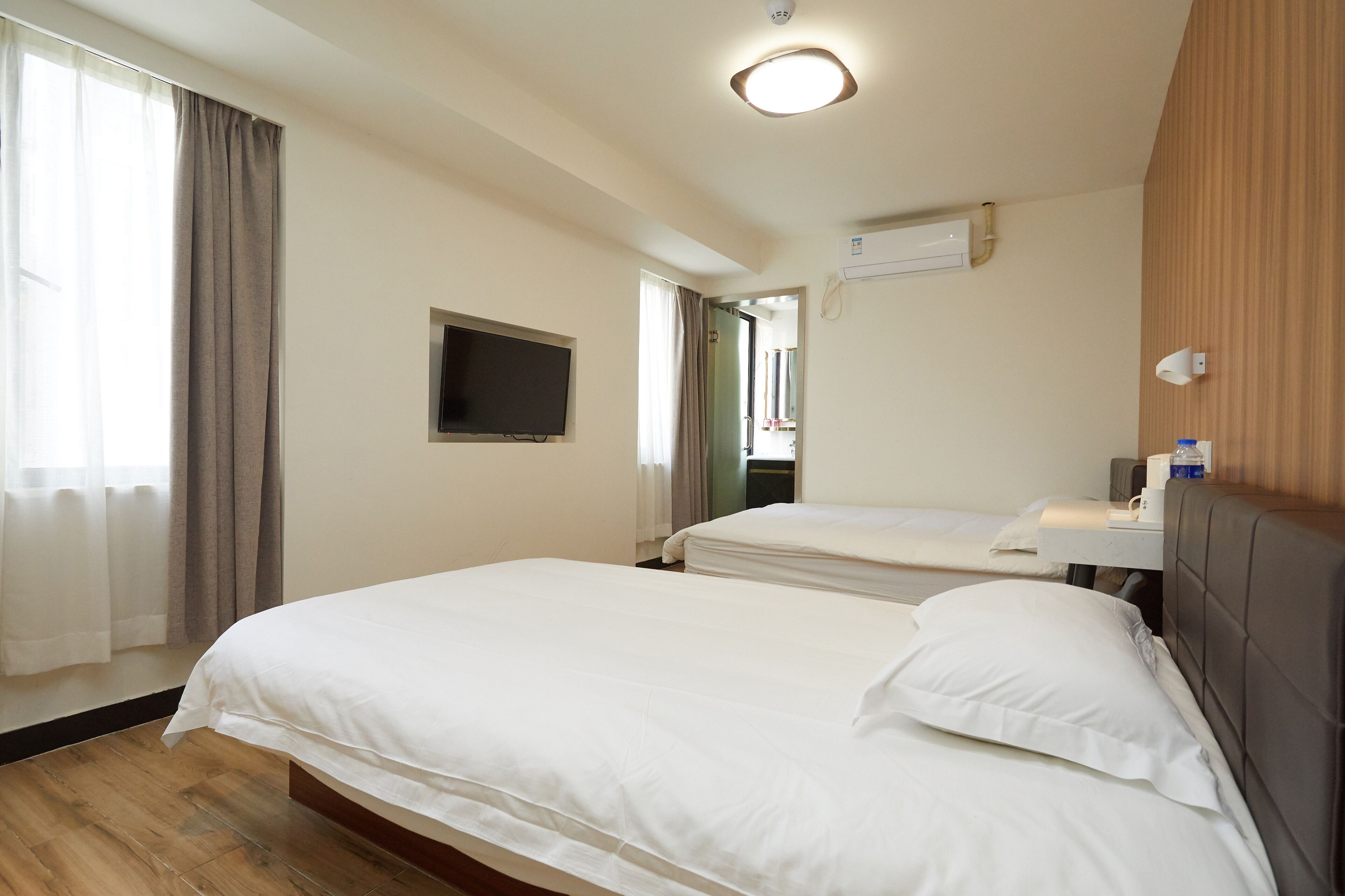 A hotel room with two beds, a flat-screen TV, an air conditioning unit, and a window with curtains.