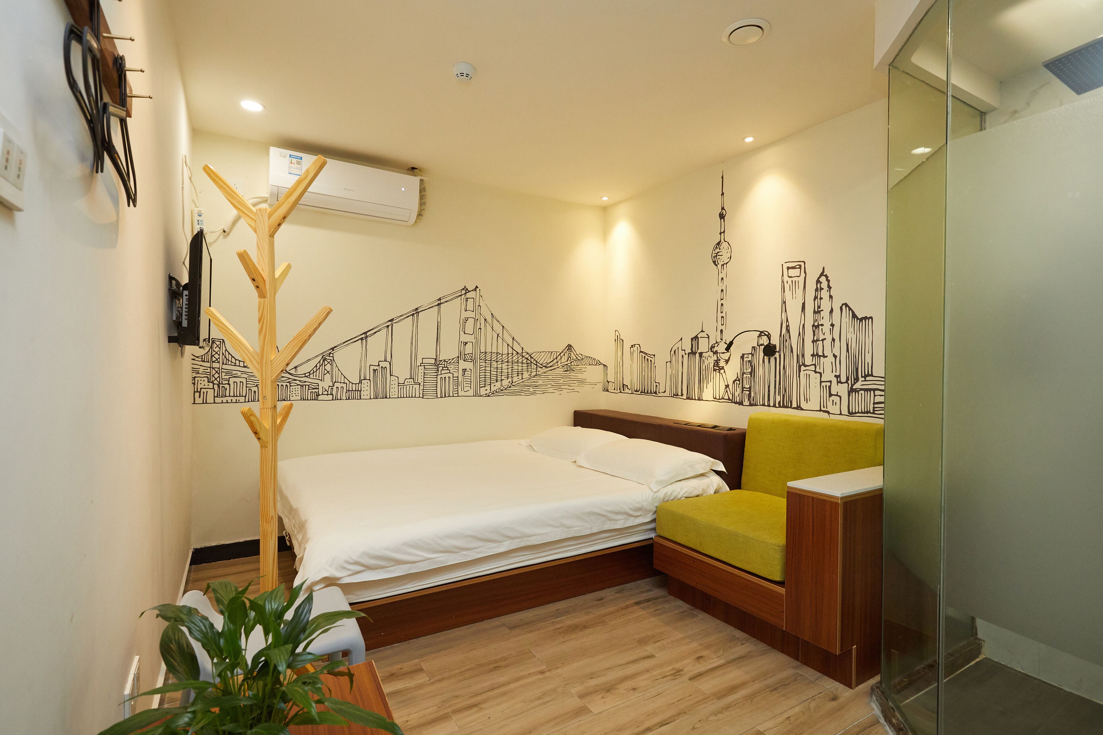 A modern hotel room with a bed, a seating area, a cityscape mural, and a plant.