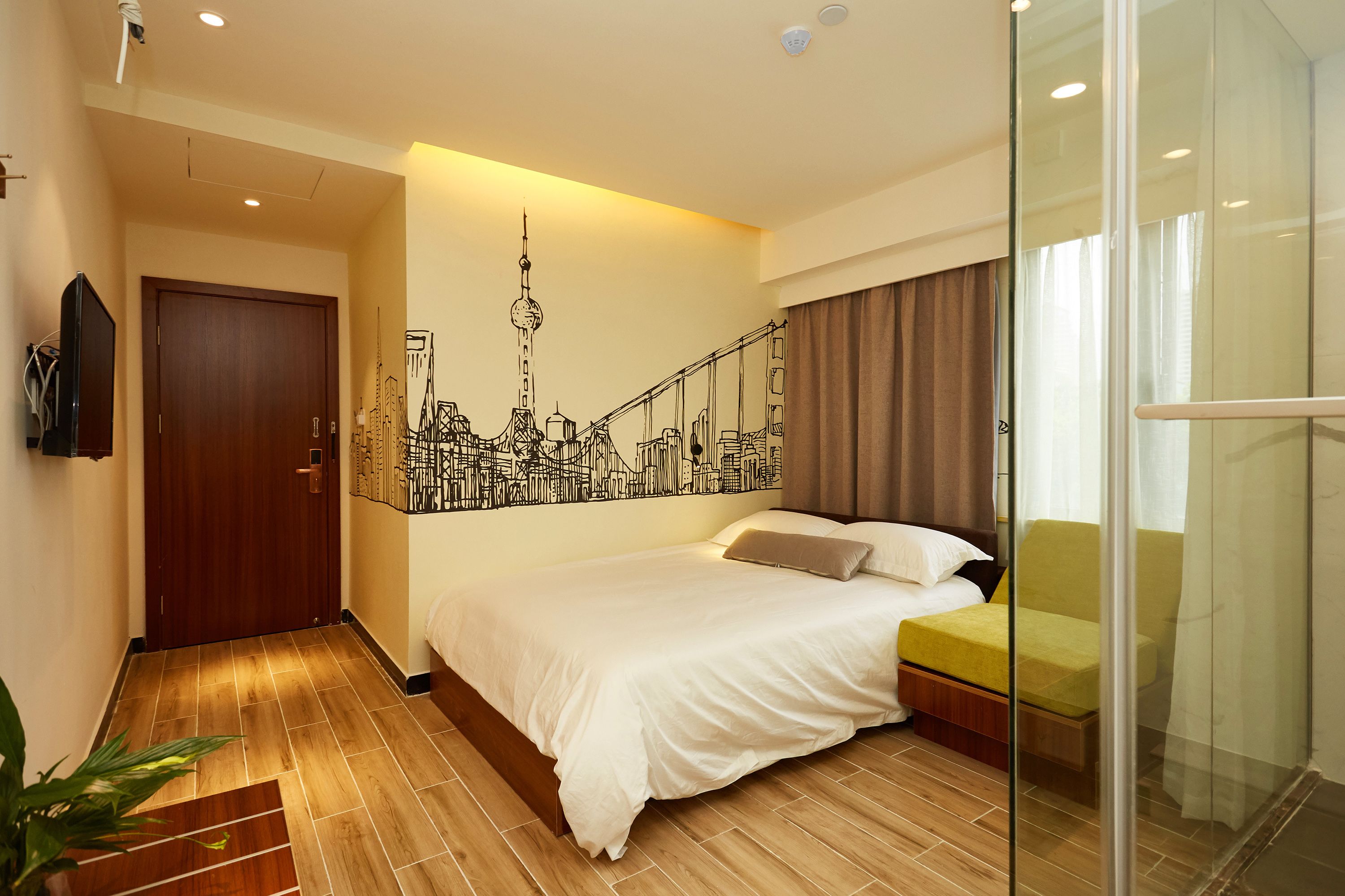 Une chambre d’hôtel avec un lit, une fresque représentant un paysage urbain, une cloison en verre et un plancher en bois.