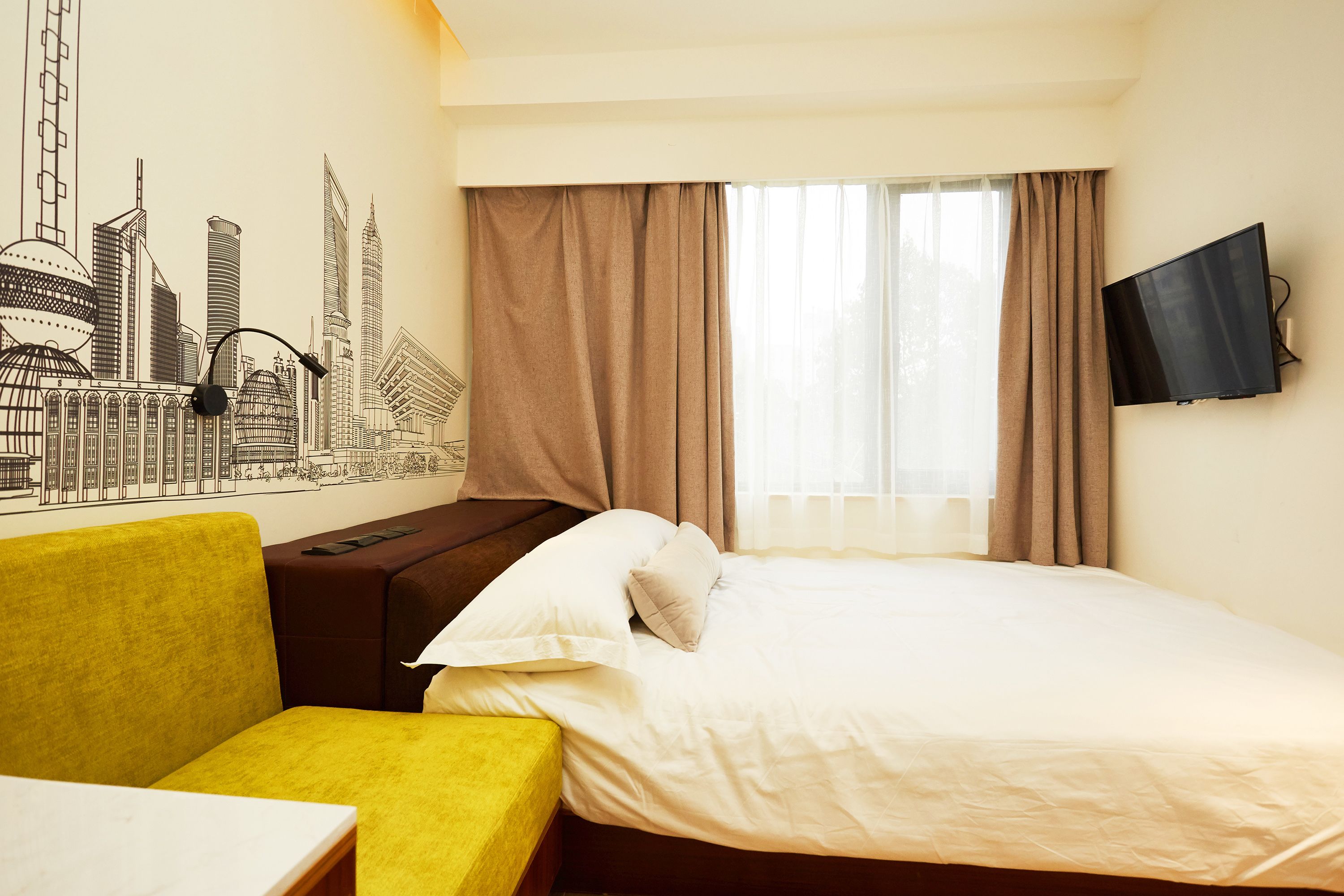 Habitación de hotel con cama, un sillón amarillo, un televisor de pantalla plana y un mural de paisaje urbano en la pared.