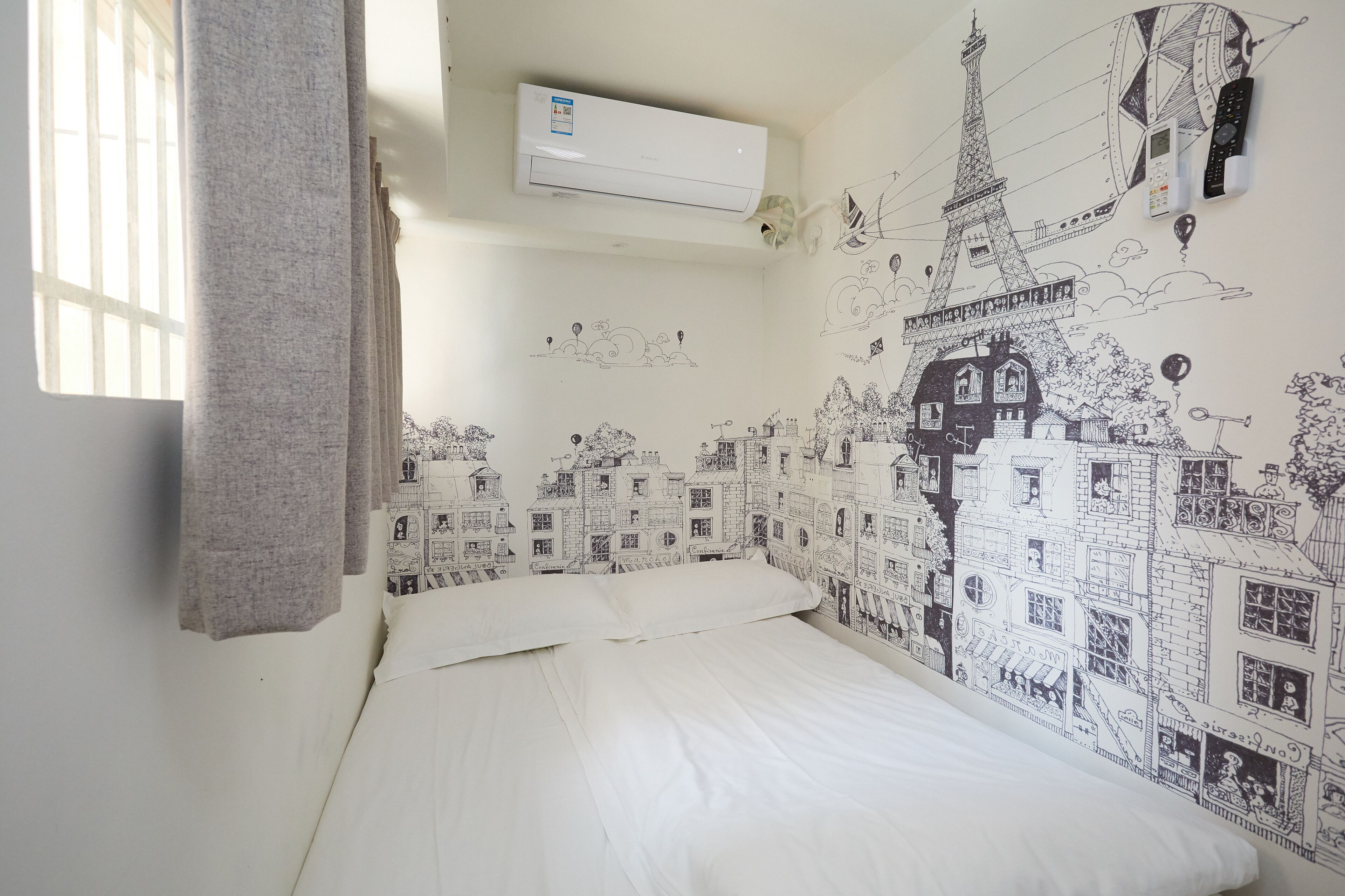 Une chambre avec un lit, du linge de lit blanc, un climatiseur et un mur orné d’une fresque représentant un paysage urbain.