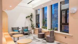 Un área de estar moderna con asientos y mesas, ventanas amplias con vistas a la ciudad y plantas de interior.