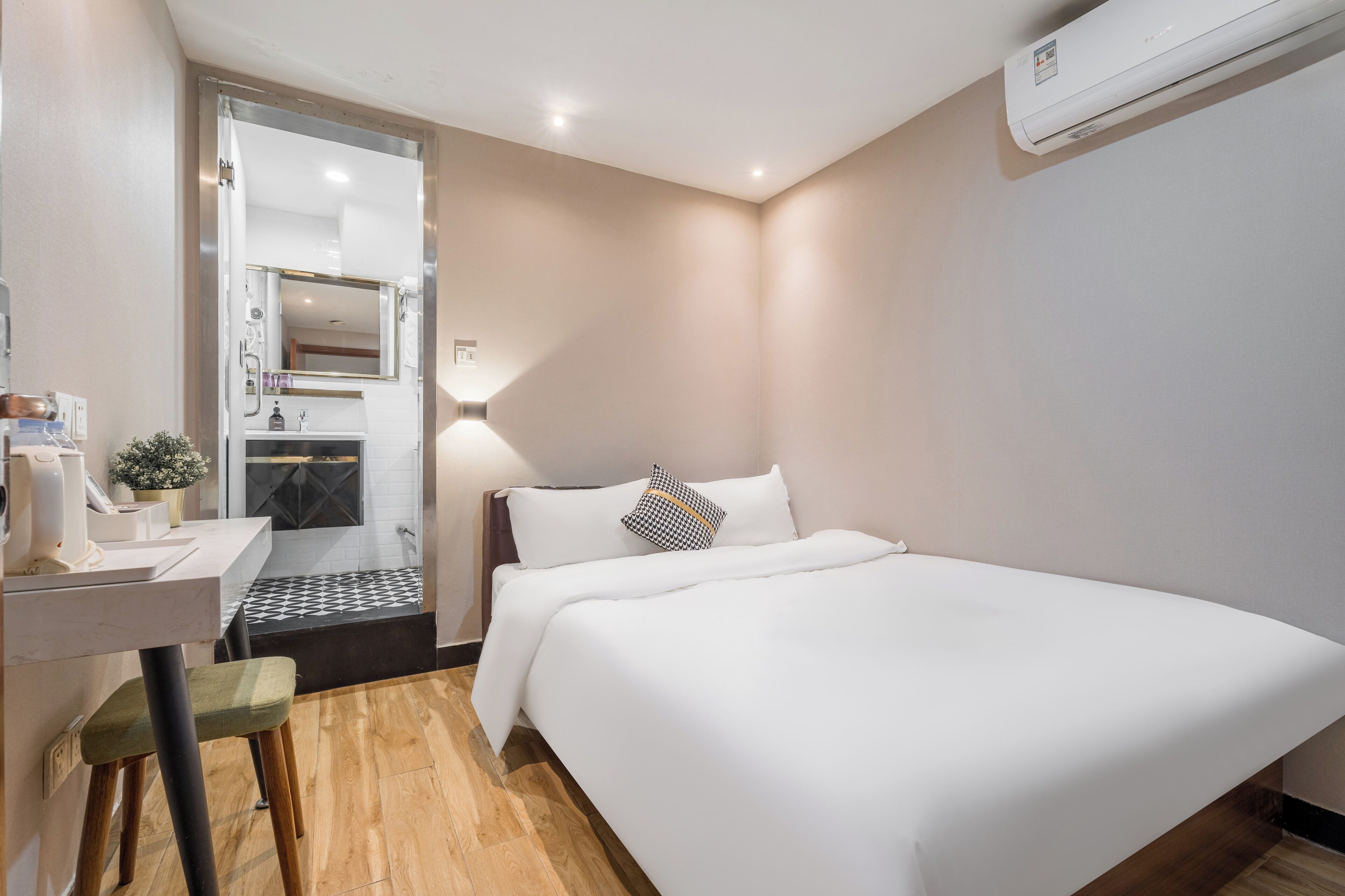 A hotel room with a bed, a desk with a chair, a mirror, and an air conditioning unit.