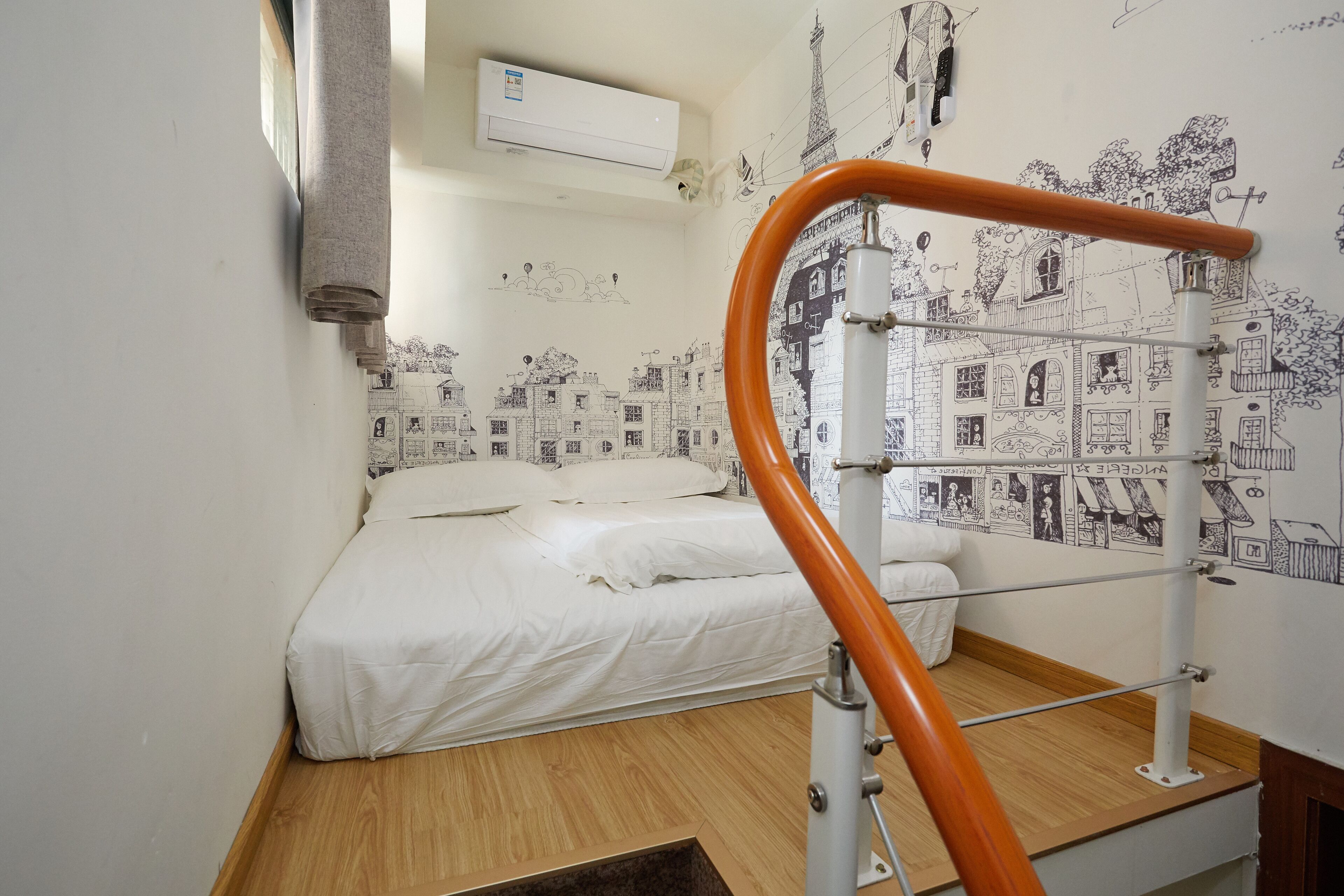 A bedroom with a bed, a wooden floor, a white wall with a cityscape mural, and an air conditioning unit.