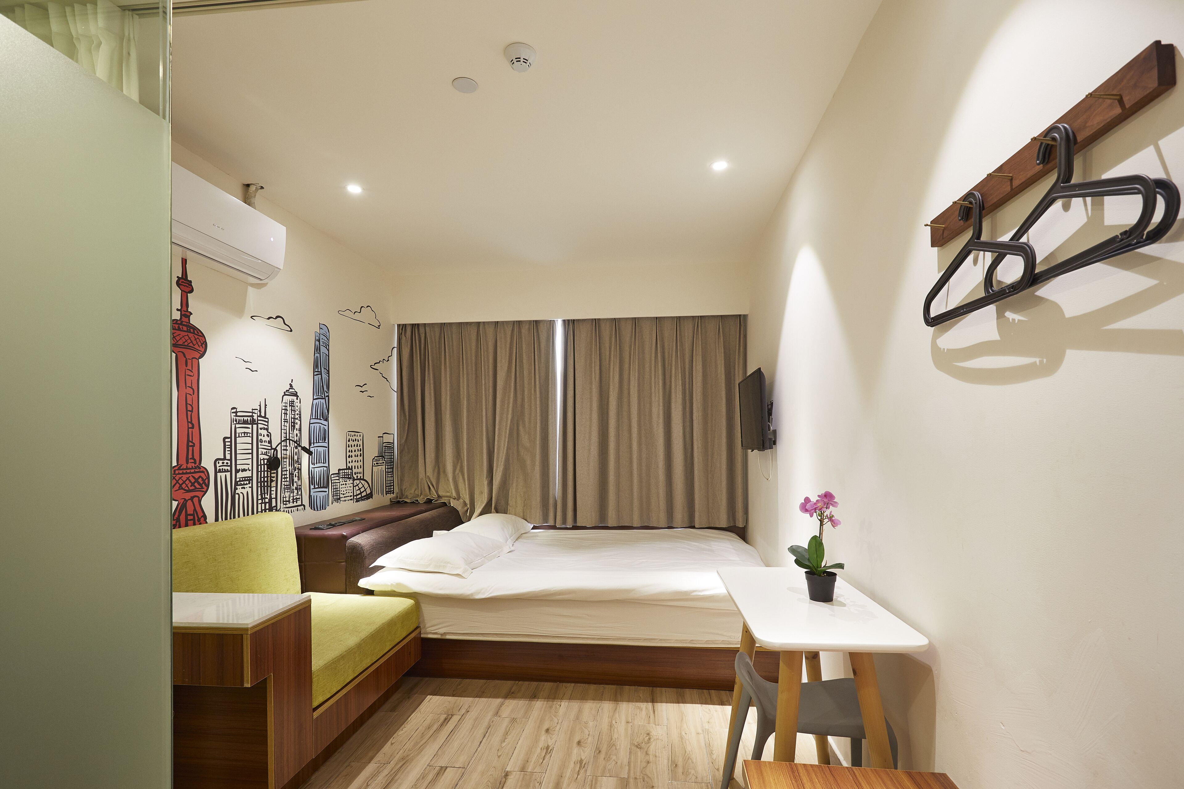 Une chambre d’hôtel de taille réduite, comprenant un lit, une petite table, une chaise, un canapé, un téléviseur et un porte-manteau fixé au mur.