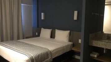 Una habitación de hotel con una cama grande, una mesita de noche, un tocador, un baño con lavamanos y espejo, y una ventana con cortinas.