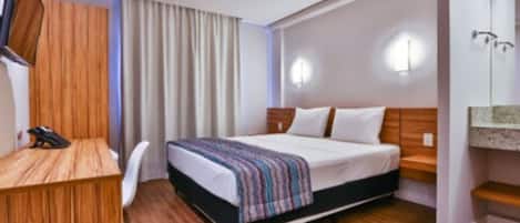 Quarto de hotel com uma cama, uma escrivaninha, uma cadeira, uma televisão e zona de banheiro.