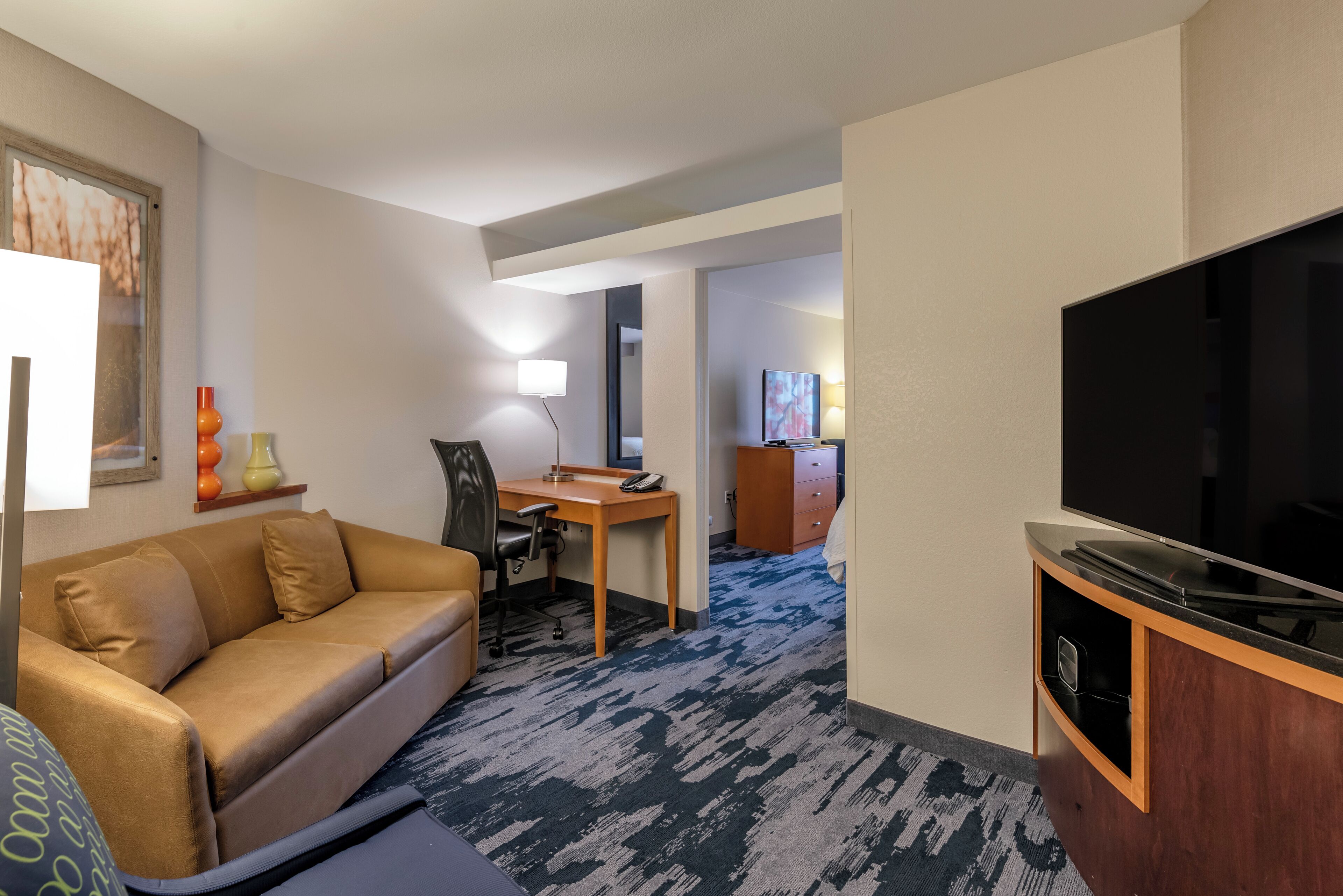 A hotel room with a sofa, desk, chair, TV, and a lamp.