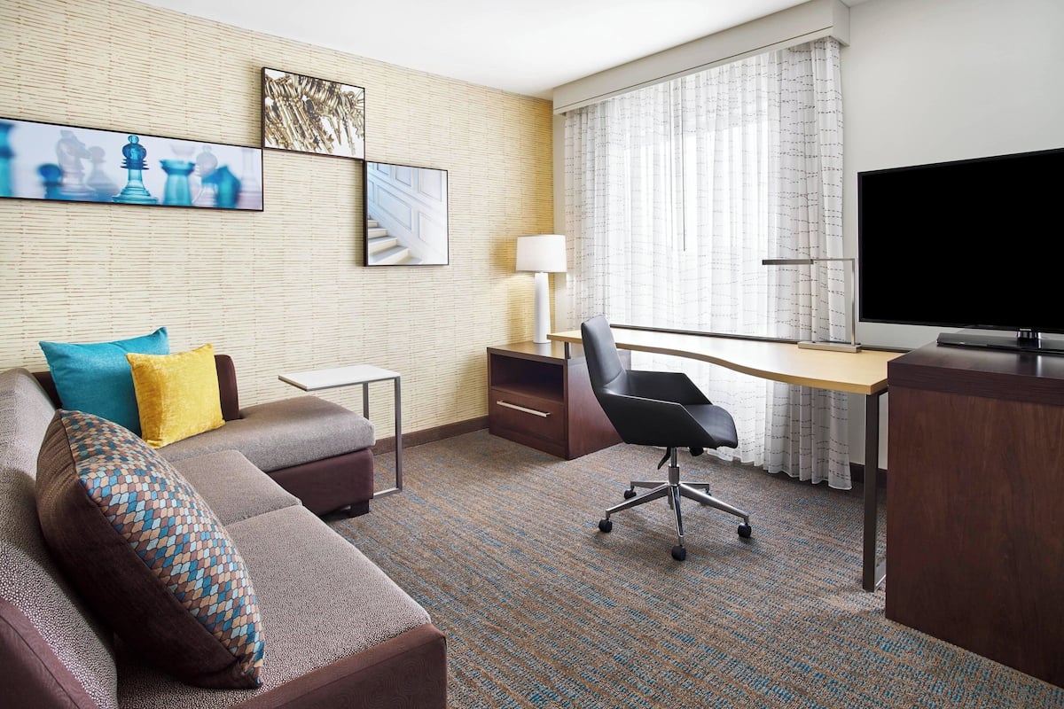 Une chambre d’hôtel équipée d’un canapé, d’un bureau, d’une chaise, d’une télévision et décorée avec des œuvres d’art aux murs.